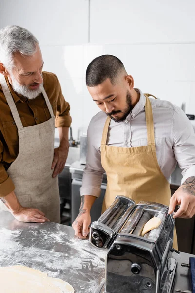 Chef asiático trabajando con la máquina de hacer pasta y pasta cerca de colega en la cocina - foto de stock