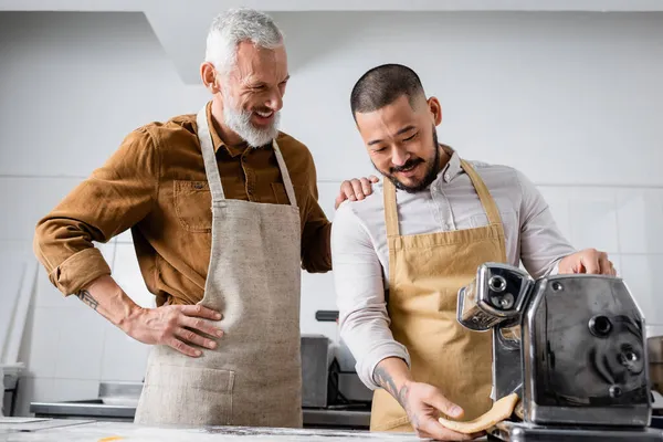 Chef sonriente en delantal de pie cerca de un colega asiático sosteniendo masa cerca de la máquina de hacer pasta en la cocina - foto de stock