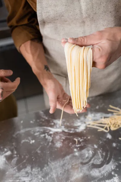Cropped view of chef holding raw spaghetti near colleague and flour on table in kitchen — Stock Photo