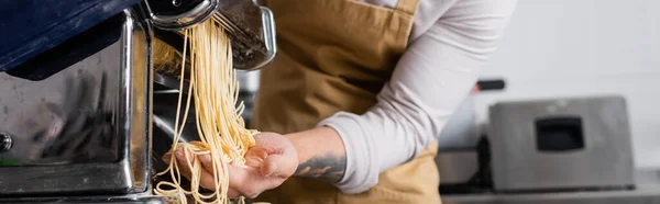 Cropped view of tattooed chef making spaghetti on pasta maker machine in kitchen, banner — Stock Photo