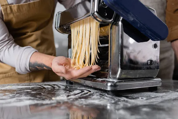 Ausgeschnittene Ansicht tätowierter Koch, der Spaghetti auf Nudelmaschine in der Nähe von Mehl auf dem Tisch macht — Stockfoto