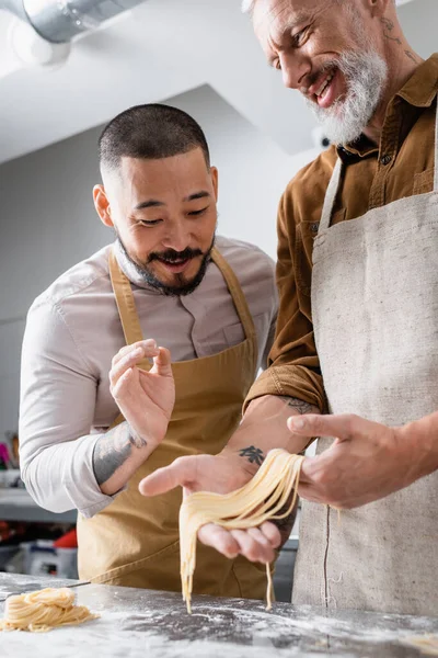 Улыбающийся азиатский шеф-повар показывает свой жест рядом с коллегой, держащим сырые спагетти на кухне. — стоковое фото
