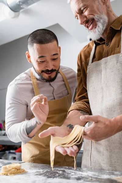 Smiling asian chef showing ok gesture near colleague holding raw spaghetti in kitchen — Stock Photo