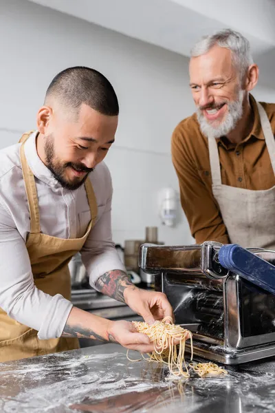 Chef asiático sosteniendo espaguetis crudos cerca de la máquina de hacer pasta y compañero borroso en la cocina - foto de stock