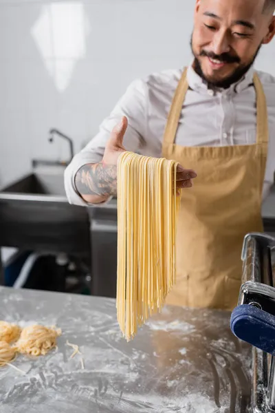 Chef asiatique flou tenant spaghettis crus près de la farine et machine à pâtes dans la cuisine — Photo de stock