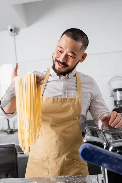 Cheerful asian chef looking at raw spaghetti near pasta maker machine in kitchen — Stock Photo