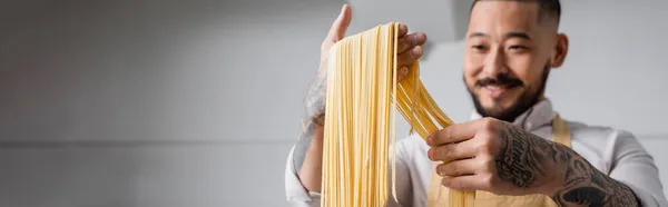 Размытый положительный азиатский шеф-повар держит сырые спагетти на кухне, баннер — стоковое фото