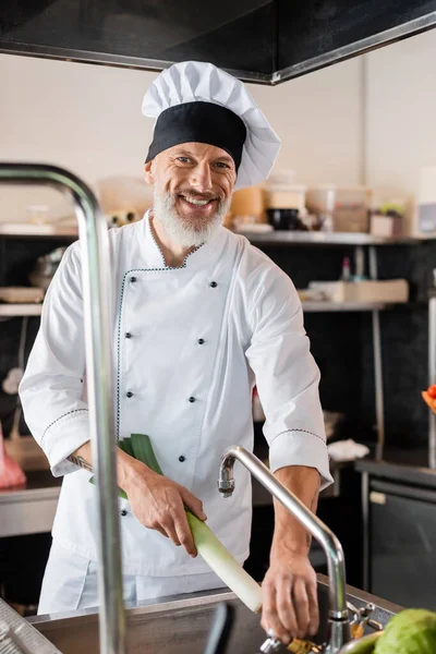 Happy mature chef holding leek near sink in kitchen — Stock Photo