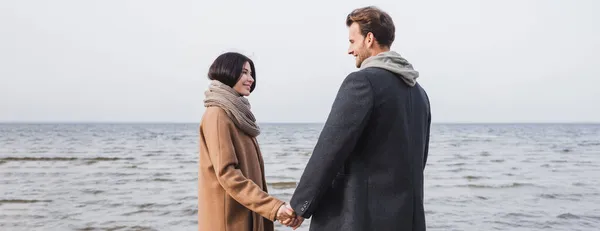 Couple heureux en manteaux d'automne tenant la main et se regardant tout en marchant près de la mer, bannière — Photo de stock
