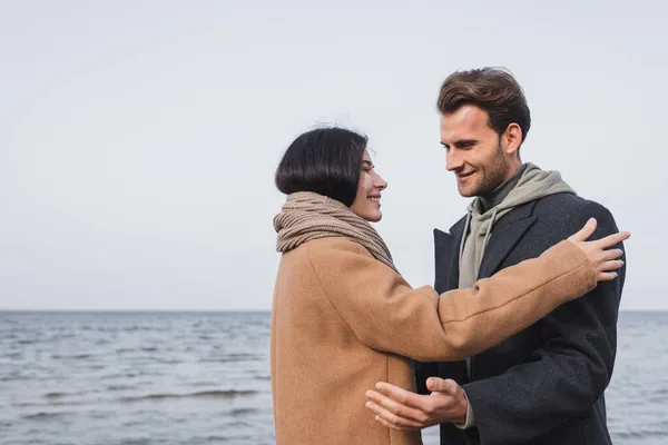 Feliz joven pareja en otoño abrigos mirando el uno al otro cerca del mar - foto de stock
