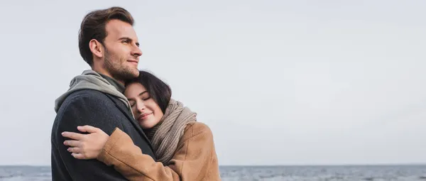 Mujer feliz con los ojos cerrados abrazando al hombre mirando lejos cerca del mar, bandera - foto de stock