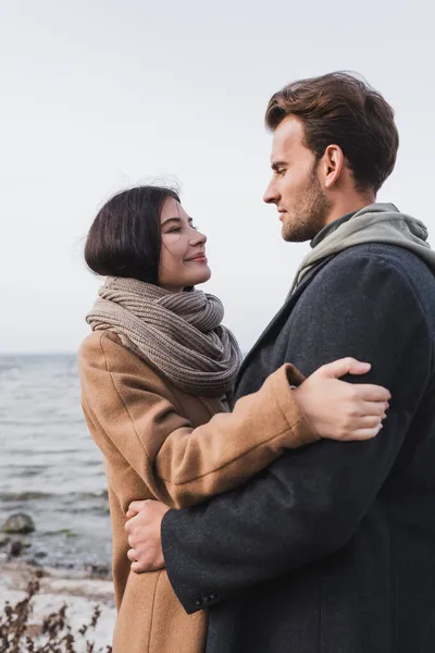 Glückliches Paar im herbstlichen Outfit, das sich am See umarmt und anschaut — Stockfoto