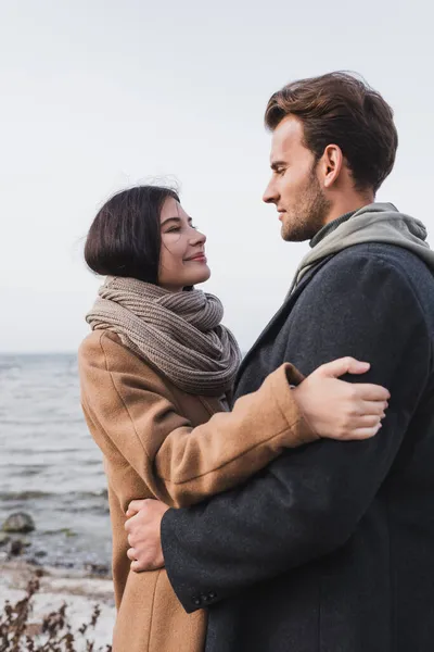 Pareja feliz en traje de otoño abrazándose y mirándose cerca del lago - foto de stock