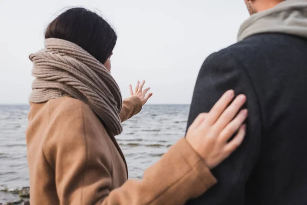 Visão traseira da mulher no casaco de outono e cachecol apontando para longe com a mão perto de homem borrado — Fotografia de Stock