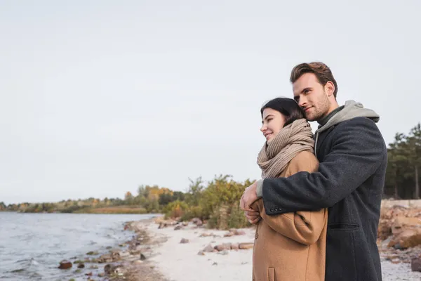 Cheerful couple in autumn outfit embracing on riverside during walk — Stock Photo