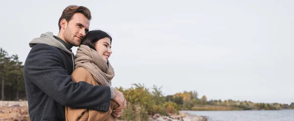 Pareja joven en traje de otoño abrazando mientras camina cerca del lago, bandera — Stock Photo