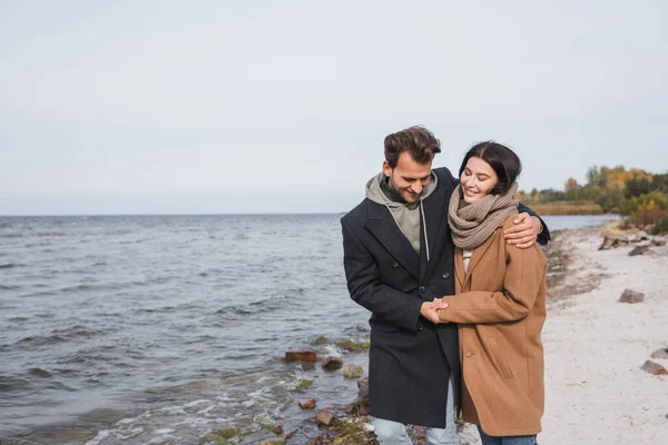 Alegre pareja en otoño abrigos abrazando anuncio cogido de la mano mientras camina a lo largo de la orilla del río - foto de stock