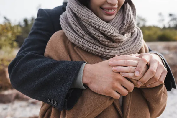 Vista parcial del hombre abrazando a la mujer sonriente en abrigo de otoño y bufanda al aire libre - foto de stock