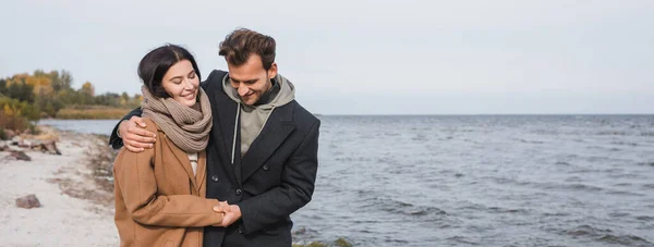 Glückliches junges Paar, das sich umarmt und Händchen hält, während es an der Küste spaziert, Banner — Stockfoto