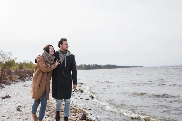 Радісна пара обіймає і дивиться в сторону під час осінньої прогулянки вздовж берега річки — стокове фото