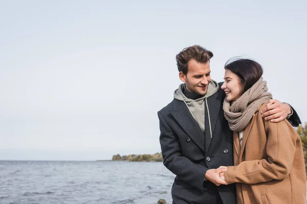 Pleased couple in autumn clothes holding hands while walking near river — Stock Photo