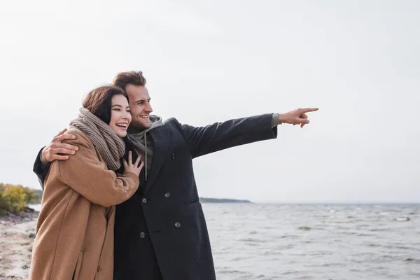 Hombre feliz señalando con el dedo mientras abraza novia cerca del mar - foto de stock