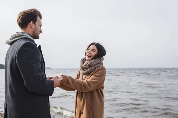 Excited woman in autumn coat holding hands with boyfriend while walking near sea — Stock Photo