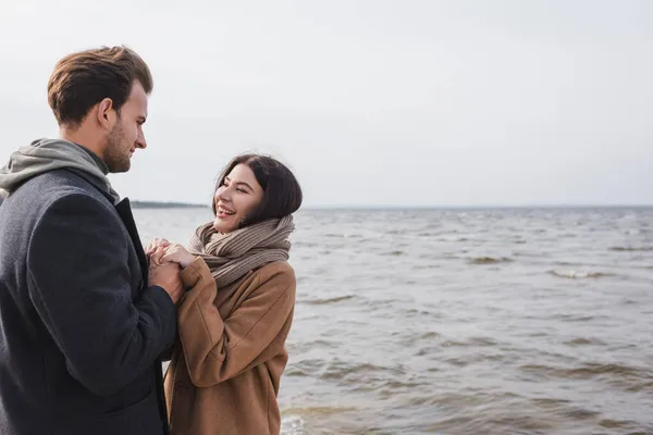 Couple joyeux tenant la main et se regardant tout en marchant près de la mer — Photo de stock