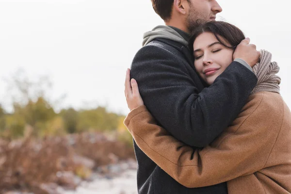 Mulher satisfeita com olhos fechados abraçando o homem no casaco de outono ao ar livre — Fotografia de Stock