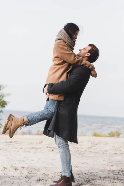 Vue de côté du jeune homme tenant femme heureuse sur le bord de la rivière — Photo de stock