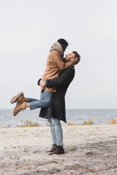 Hombre feliz en ropa de otoño sosteniendo mujer en las manos cerca del río - foto de stock