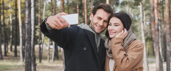 Uomo e donna sorridenti in cappotti autunnali che scattano selfie su smartphone nel parco, banner — Foto stock