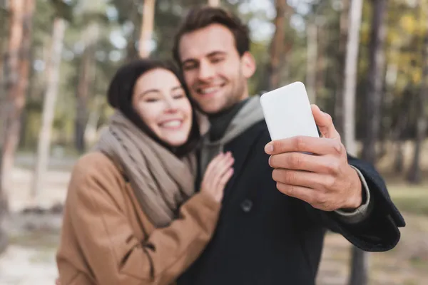 Borrosa y feliz pareja tomando selfie en el teléfono inteligente mientras camina en el bosque de otoño - foto de stock