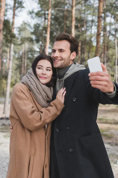 Mujer alegre abrazando novio tomando selfie en el teléfono móvil en el bosque de otoño - foto de stock