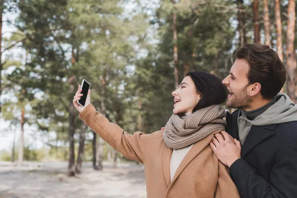 Couple excité en manteaux d'automne prenant selfie sur smartphone avec écran blanc en forêt — Photo de stock