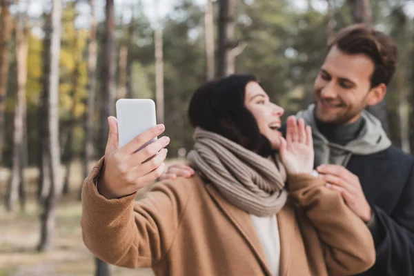 Mujer emocionada tomando selfie en el teléfono celular con novio en el bosque sobre fondo borroso - foto de stock