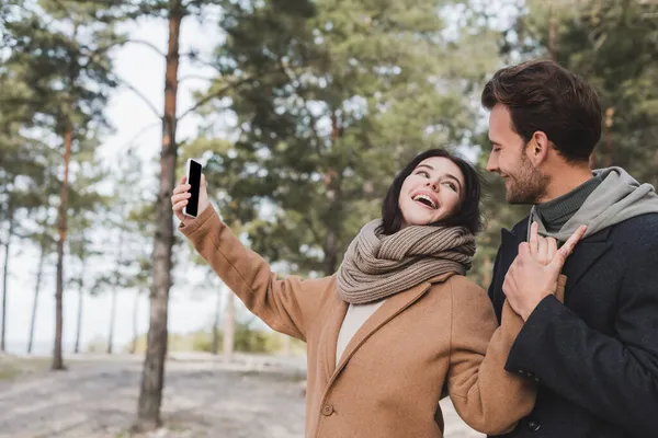 Mujer alegre en abrigo de otoño tomando selfie en el teléfono móvil con pantalla en blanco mientras camina en el parque - foto de stock