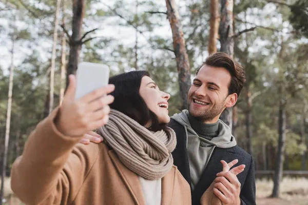 Cheerful woman taking selfie on blurred cellphone with boyfriend during walk in forest — Stock Photo