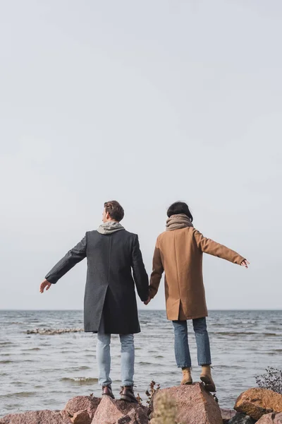 Vista trasera de pareja joven en abrigos de otoño tomados de la mano mientras están de pie en la costa del mar - foto de stock