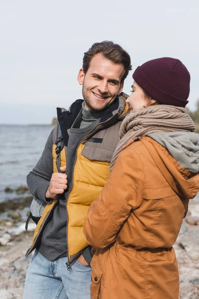 Hombre sonriente en ropa de otoño mirando a su novia mientras camina cerca del río - foto de stock