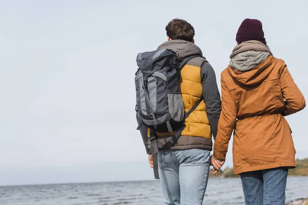 Vue arrière du couple en tenue d'automne tenant la main lors d'une randonnée près de la rivière — Photo de stock