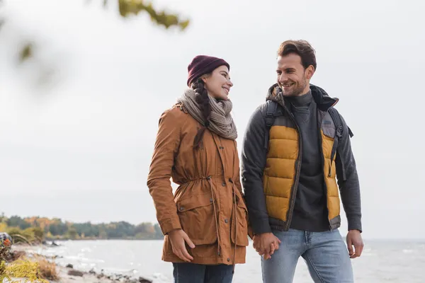 Happy young couple talking and holding hands while walking near lake — Stock Photo