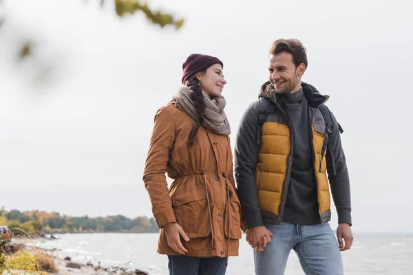 Heureux jeune couple parler et se tenant la main tout en marchant près du lac — Photo de stock