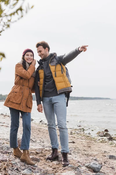 Homme pointant du doigt tout en marchant avec la femme le long de la rive du lac en automne — Photo de stock