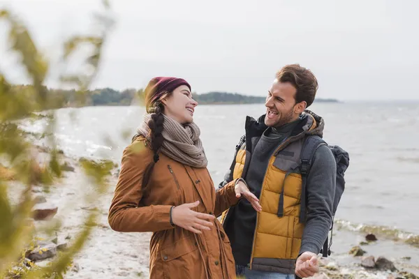 Excited couple talking and laughing while walking near river on blurred foreground — Stock Photo