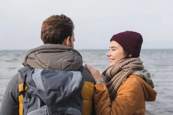 Vista posteriore dell'uomo con zaino vicino alla donna sorridente che si tocca la spalla durante la passeggiata vicino al mare — Foto stock