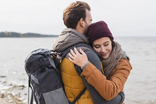 Mulher feliz com olhos fechados abraçando namorado com mochila enquanto caminhava perto do rio — Fotografia de Stock