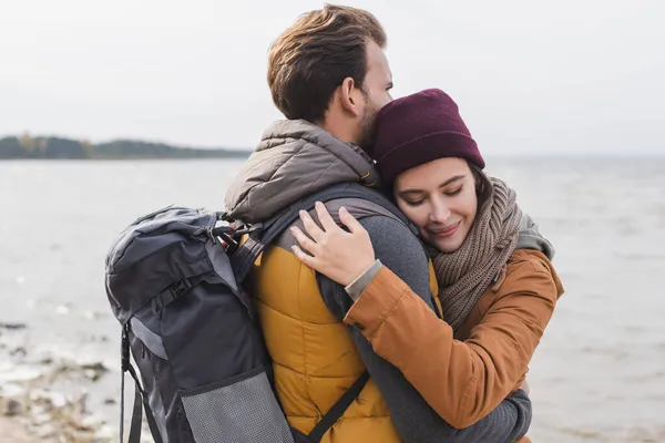 Donna felice con gli occhi chiusi abbracciare fidanzato con lo zaino mentre si cammina vicino al fiume — Foto stock