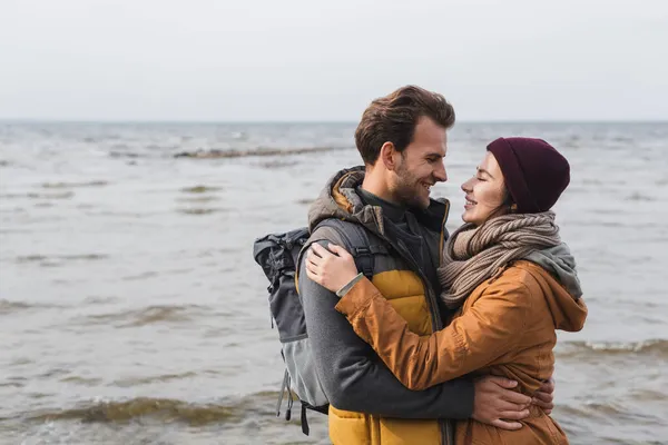 Pareja joven en traje de otoño abrazándose y mirándose cerca del mar - foto de stock