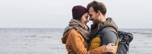 Vista lateral de la feliz pareja en traje de otoño abrazando con los ojos cerrados cerca del río, bandera - foto de stock